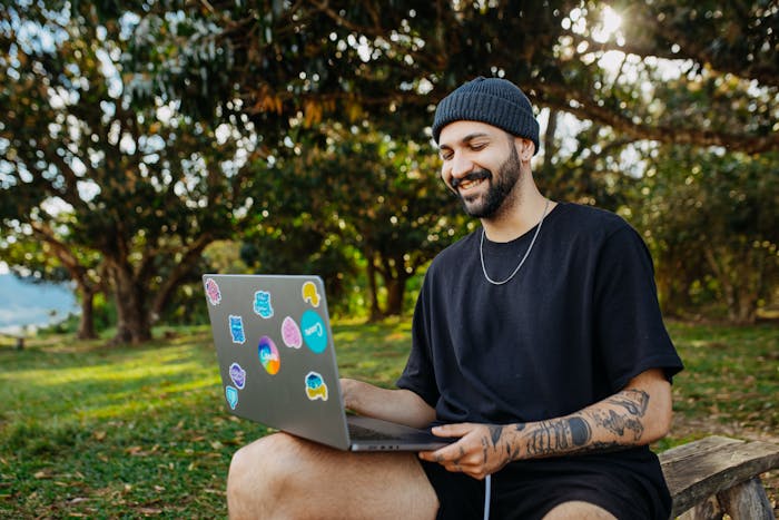 A smiling man with tattoos working on a laptop outdoors in a sunny park setting.