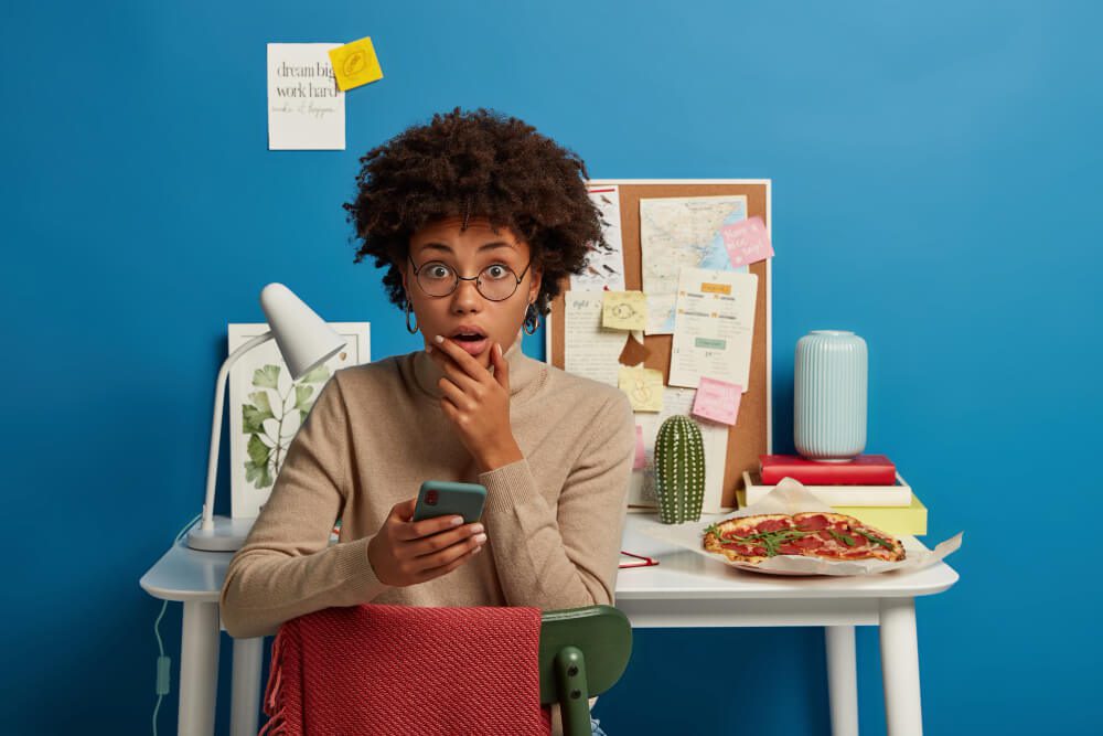 A person with curly hair and glasses looks surprised while holding a smartphone, perhaps after checking their X handle, sitting at a desk with books, a lamp, cactus, pizza, and a corkboard with notes. Uploaded on aifreeforever.com