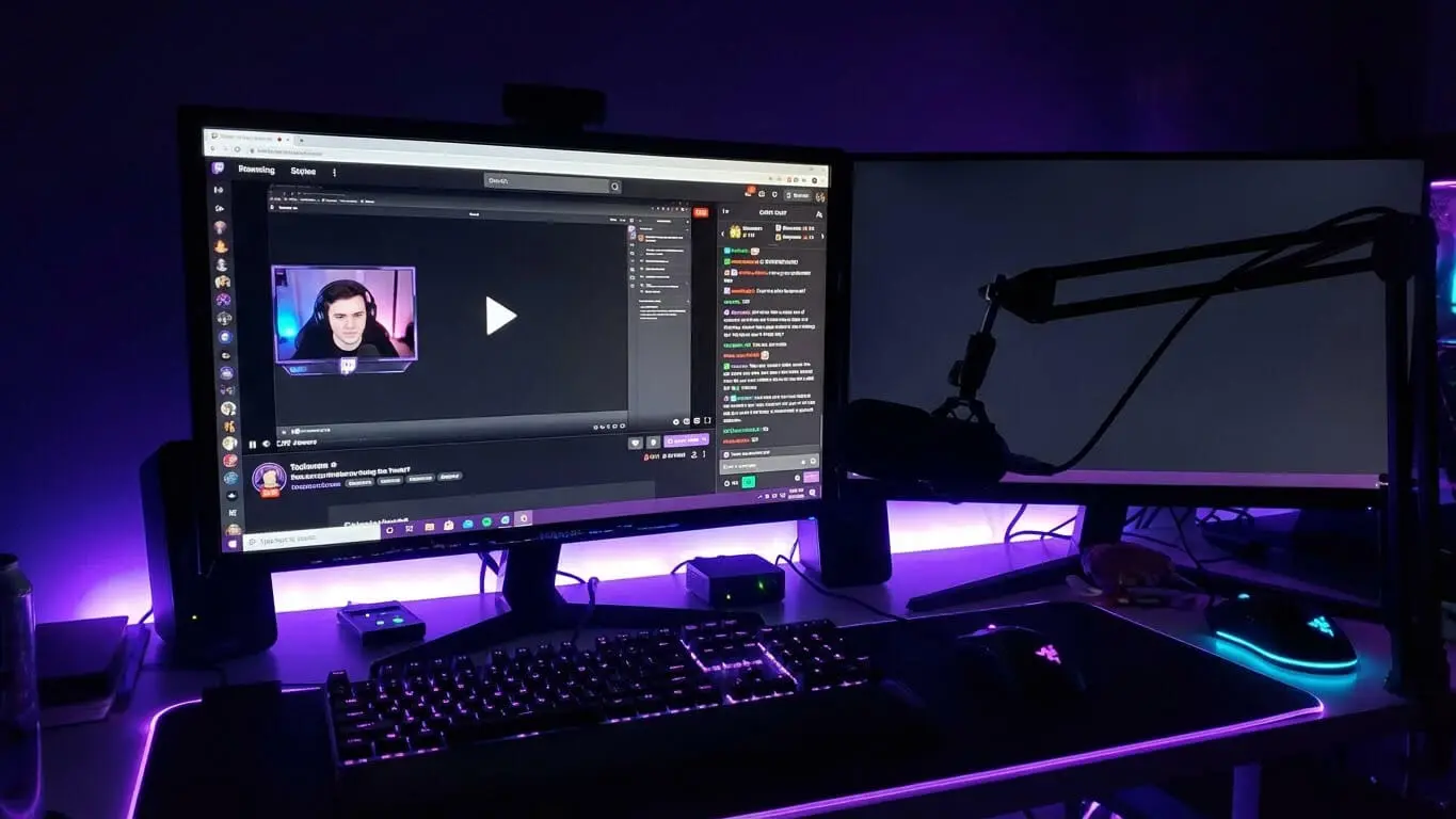 A computer setup with dual monitors—one showing a livestream—glows with purple LED lights. On the desk sit a microphone, keyboard, and mouse, perfect for brainstorming Streamer Names or using the Twitch Name Generator from Namelix. Uploaded on aifreeforever.com