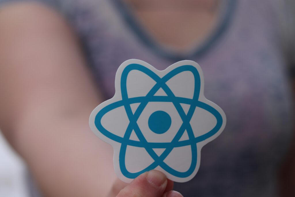 A person holds a sticker featuring the blue React logo, which resembles an atom, with a blurred background—perfect for showcasing your favorite AI tools or social media AI tools. .Uploaded on aifreeforever.com