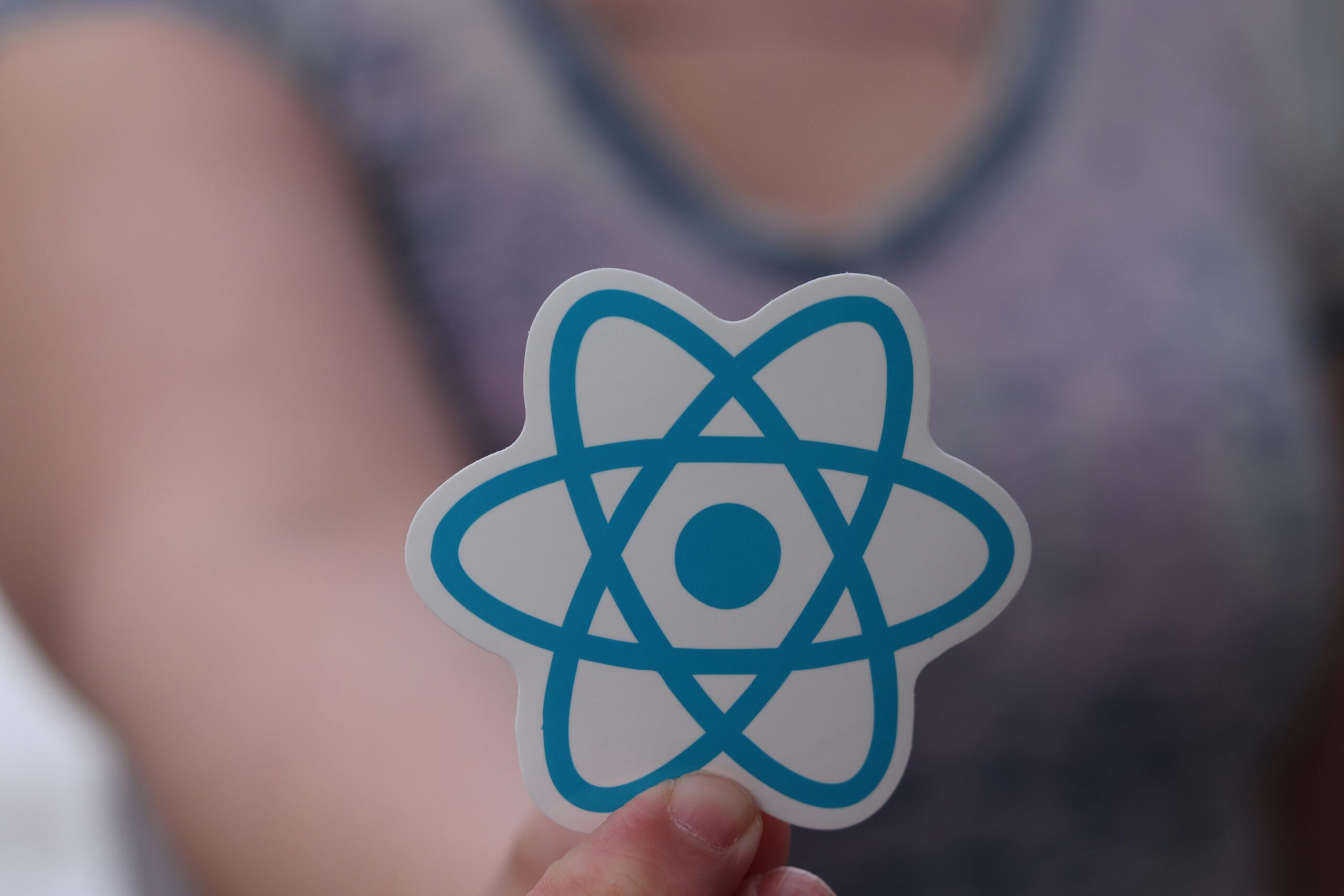 A person holds a sticker featuring the blue React logo, which resembles an atom, with a blurred background—perfect for showcasing your favorite AI tools or social media AI tools. .Uploaded on aifreeforever.com